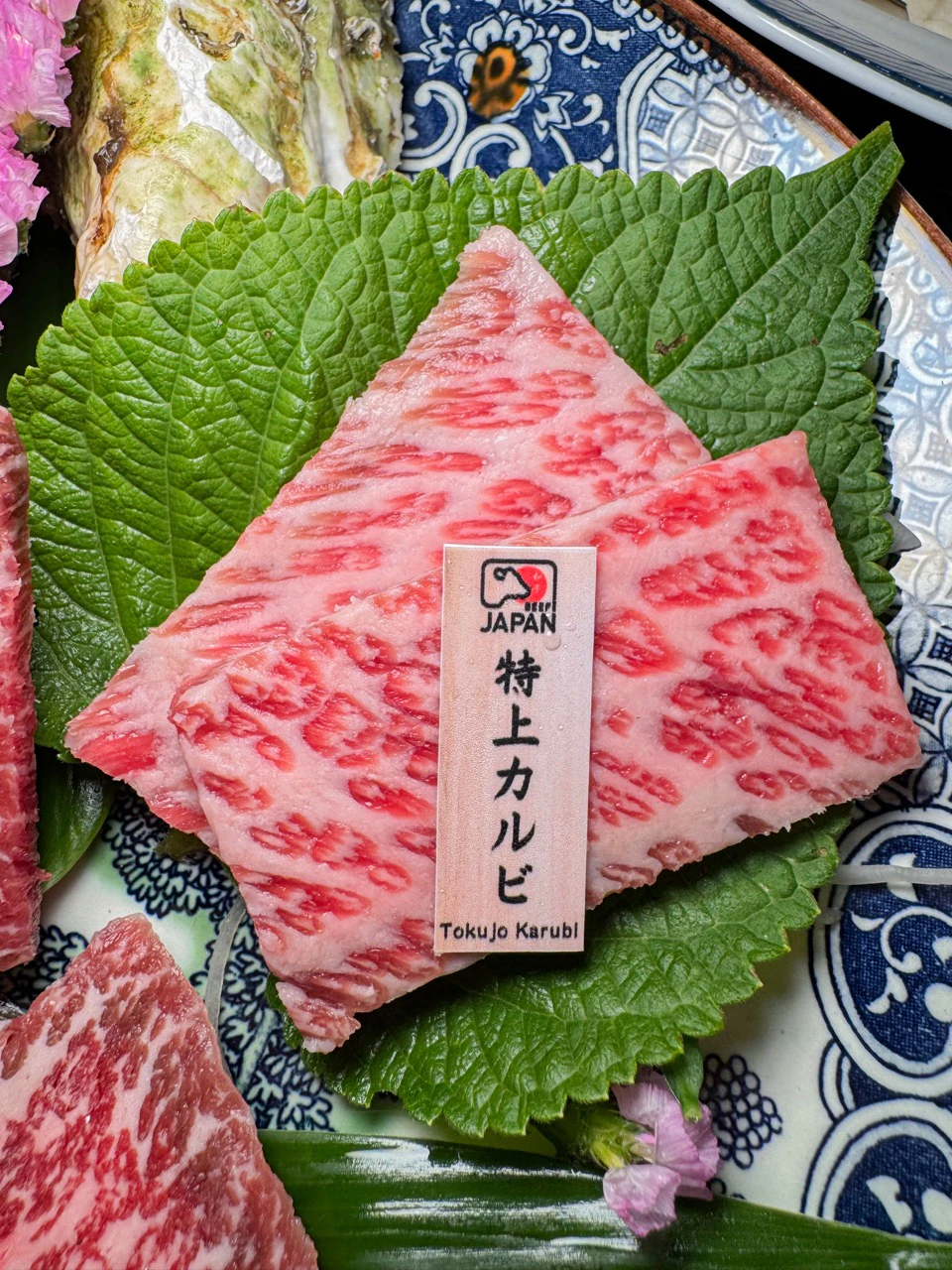 Two marbled pieces of raw meat on green leaves with a label reading "Japan Tokujou Karubi" placed on top.