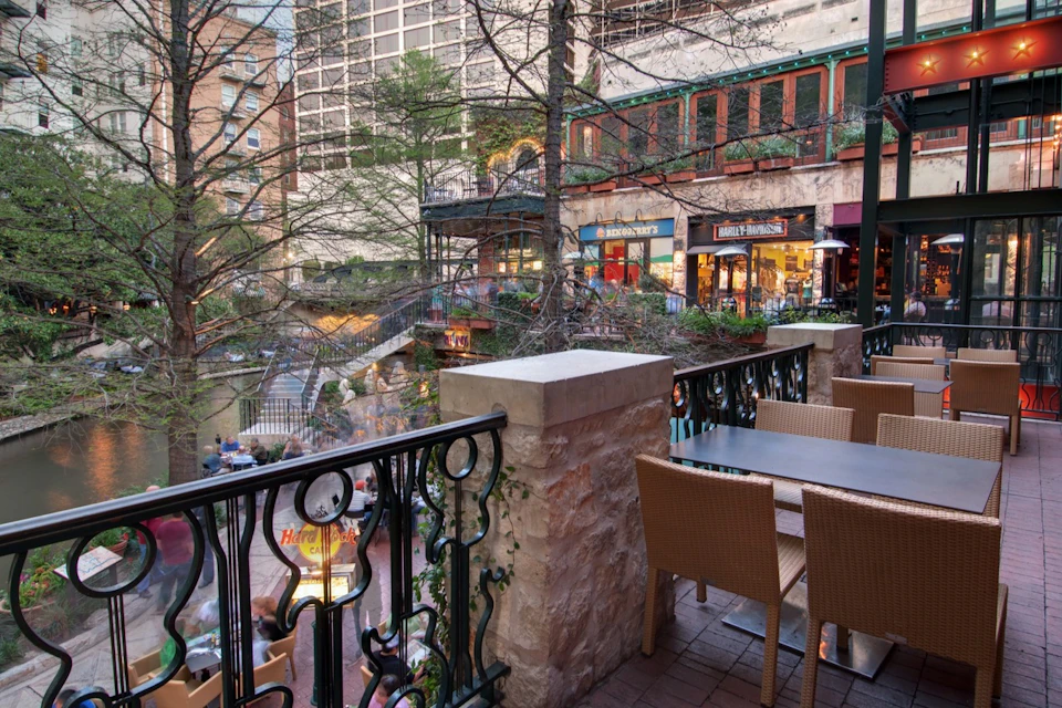Outdoor café seating on a balcony overlooks a shopping area with trees, railings, and storefronts in an urban setting.