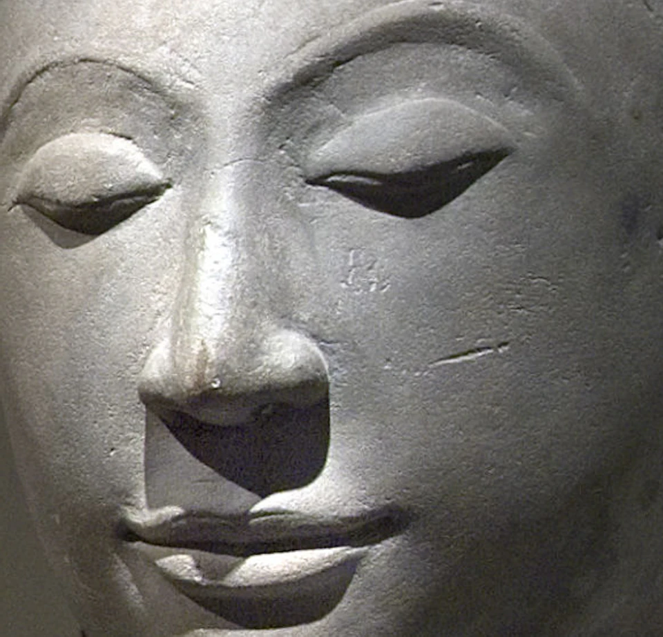 Museum gallery with sculptures and artifacts displayed on pedestals. Foreground features a large stone head and a wooden cabinet.