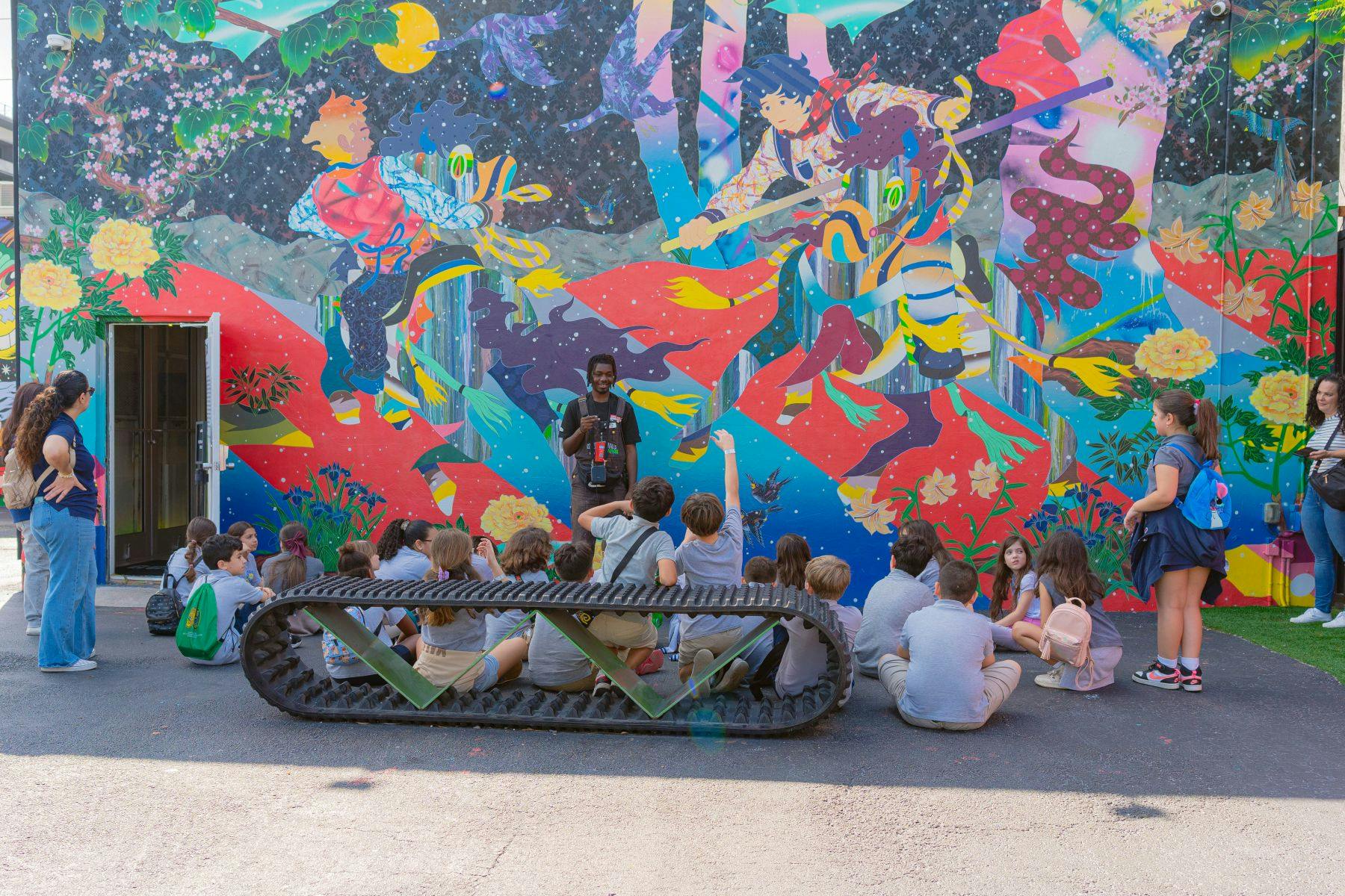 一群孩子坐在地上，在一幅充满活力、色彩缤纷的壁画前听导游讲解。