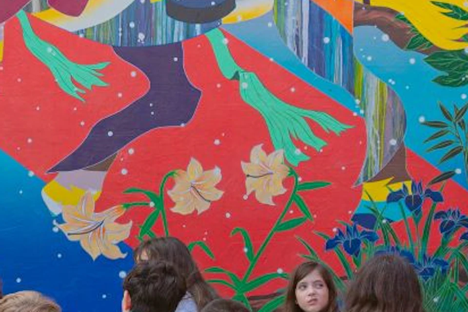A group of children sit on the ground listening to a guide in front of a vibrant, colorful mural.