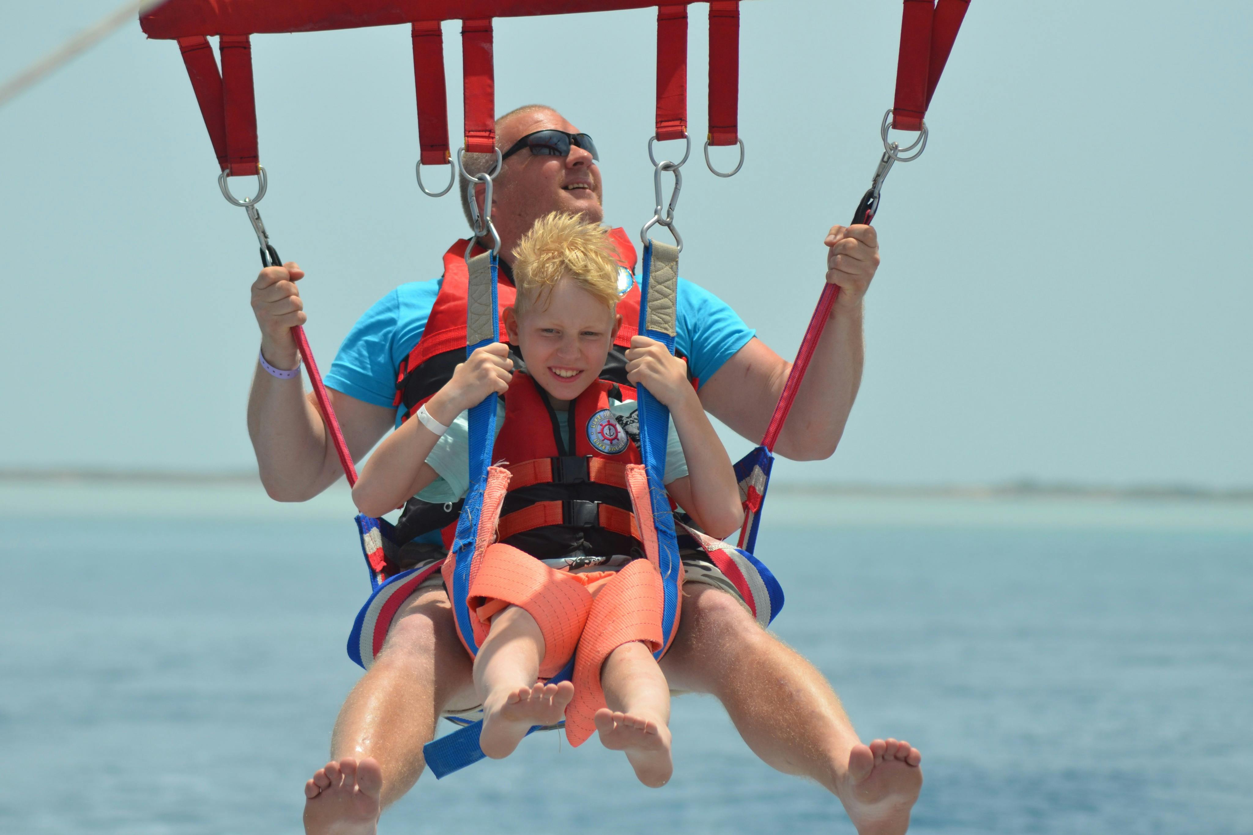 A man and a child parasailing, wearing life jackets and harnesses, with a clear blue sky and ocean in the background.