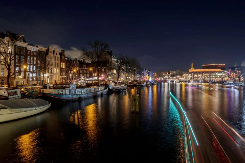 City canal at night, illuminated buildings and boats reflecting on water, with light trails along the water's edge.