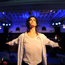 En vaxstaty med utsträckta armar framför en scen med två stora skärmar och blå belysning i bakgrunden.