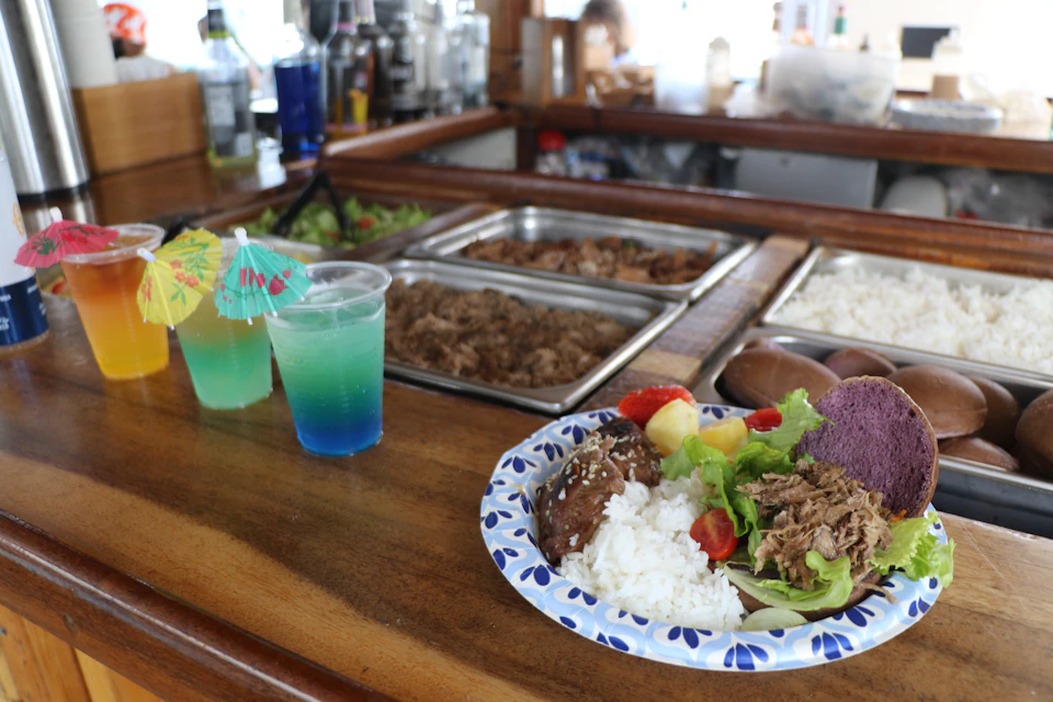 A plate of food with rice, meat, and vegetables on a wooden counter next to two colorful drinks and a food serving station.