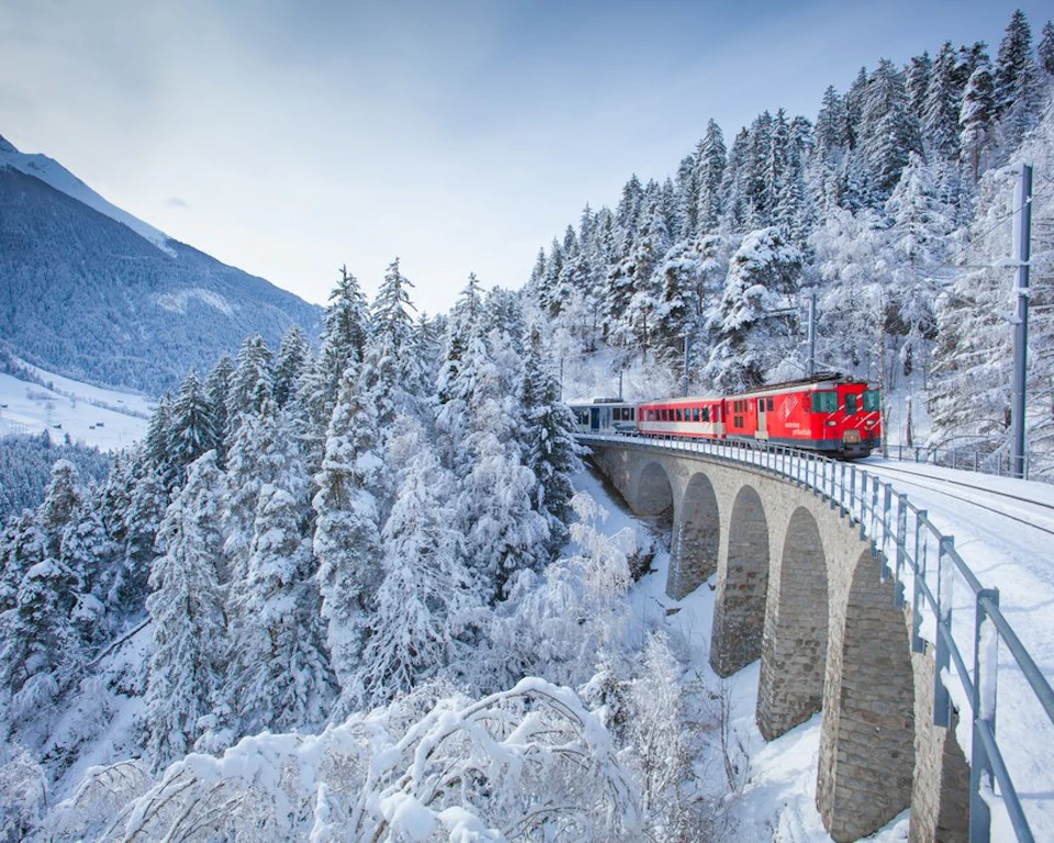 A red train travels over a stone bridge through a snowy forested landscape with mountains in the background.