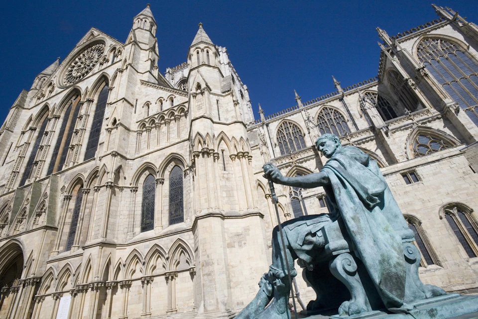 Statue of a seated figure with a sword in front of a large, ornate Gothic cathedral under a clear blue sky.