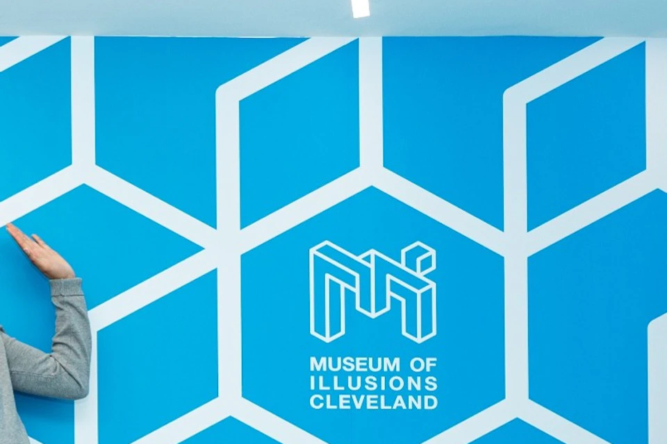 Four people stand in a room with a blue geometric pattern wall, labeled "Museum of Illusions Cleveland."