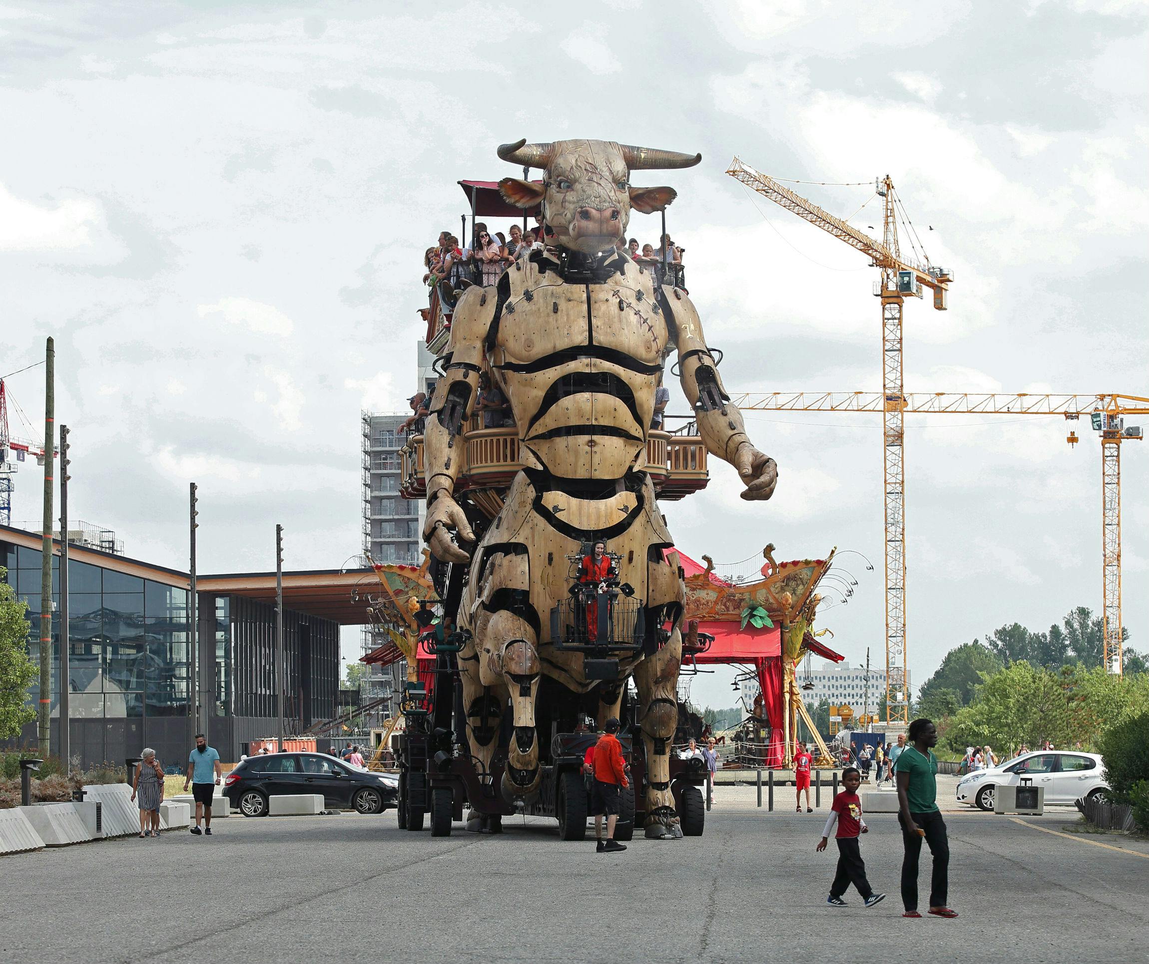 背上有人的大型机械公牛穿过街道。 后台有围观者、起重机、建筑物和汽车。