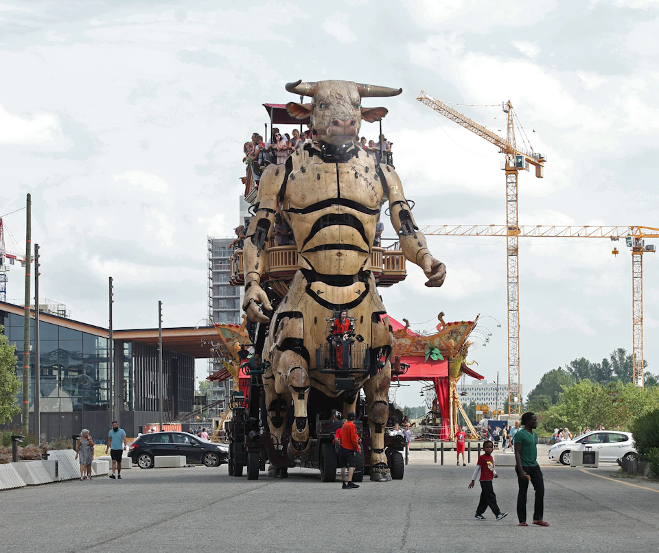 Large mechanical bull with people on its back moves through a street. Onlookers, cranes, buildings, and cars in the background.