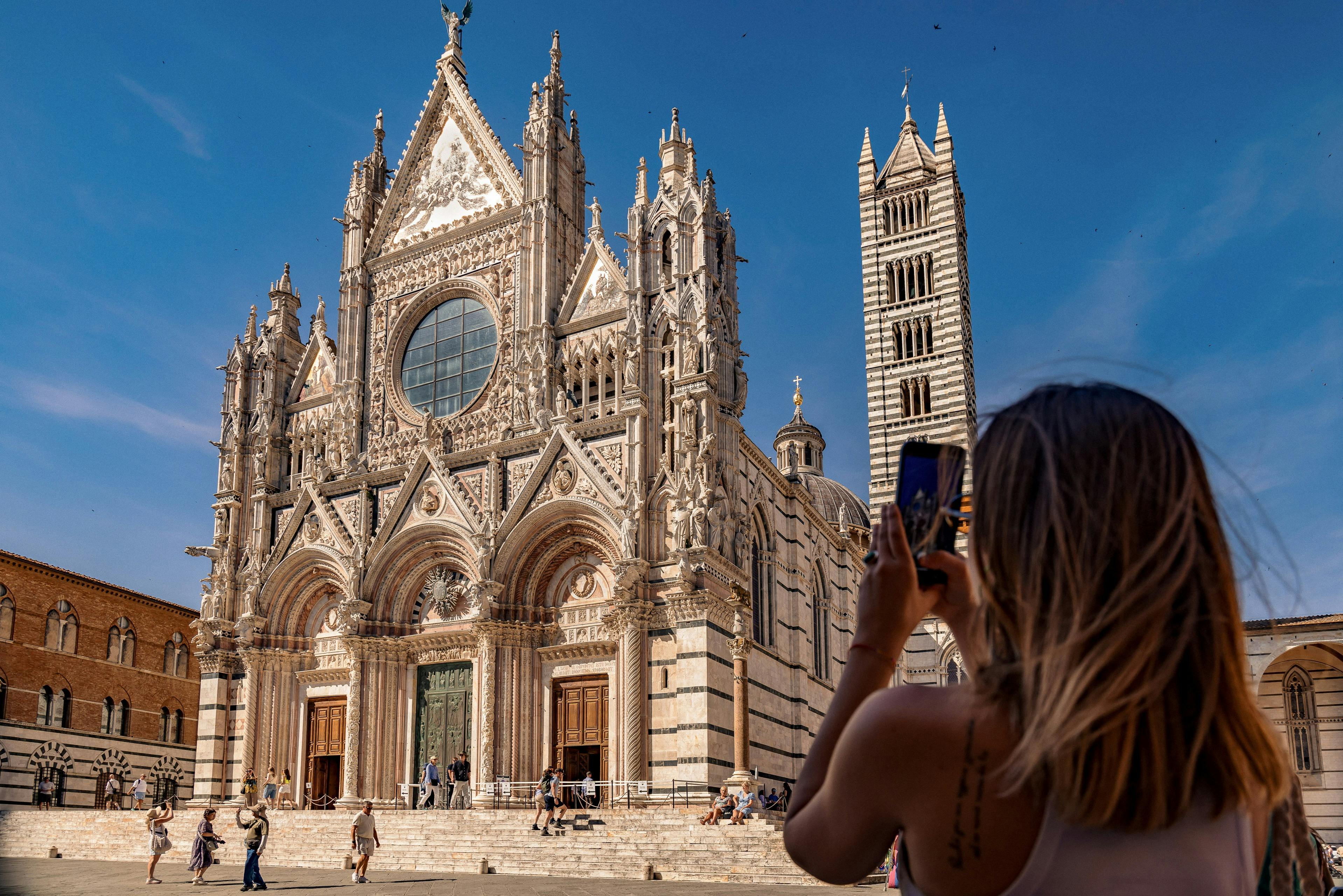 Una persona che scatta una foto alla facciata decorata di una cattedrale con un alto campanile sotto un cielo azzurro e limpido.