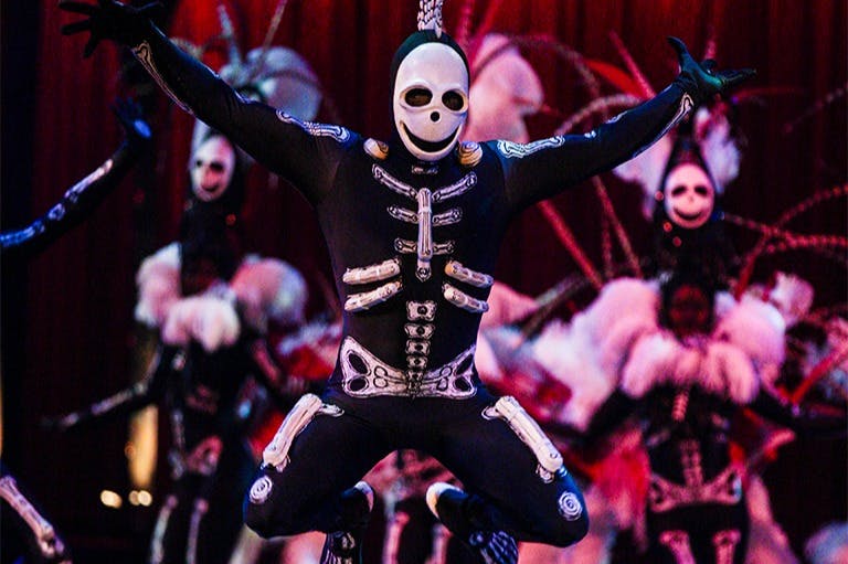 Una intèrpret amb un disfressa d'esquelet ballant a l'escenari, amb intèrprets disfressats similars i una cortina vermella al fons.