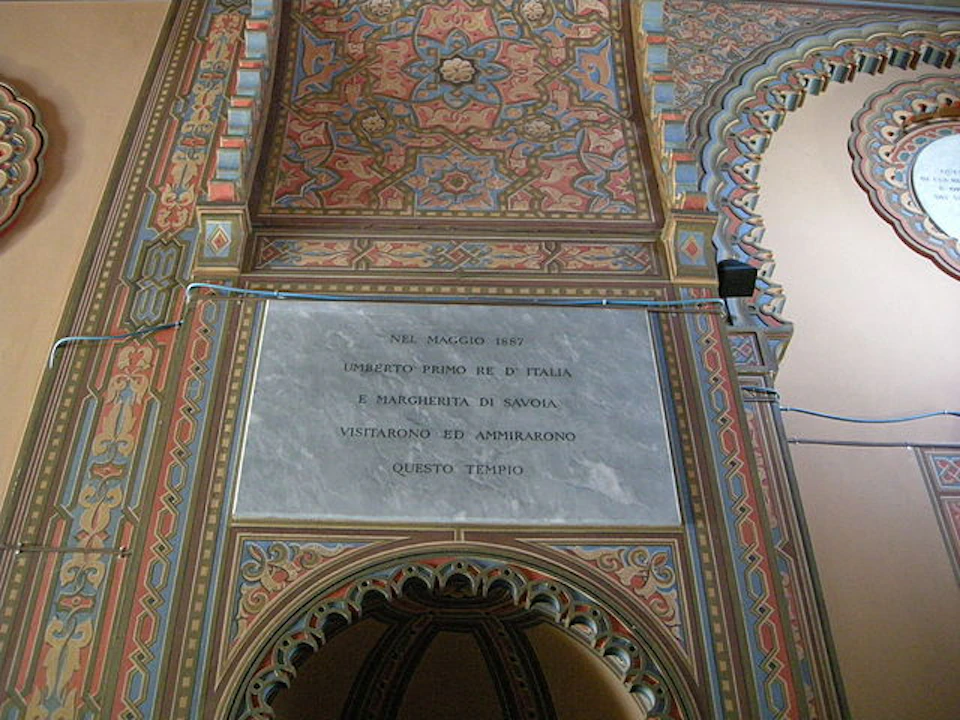 An ornate, arched wall with detailed patterns and Italian text on a marble plaque, commemorating a visit in May 1887.