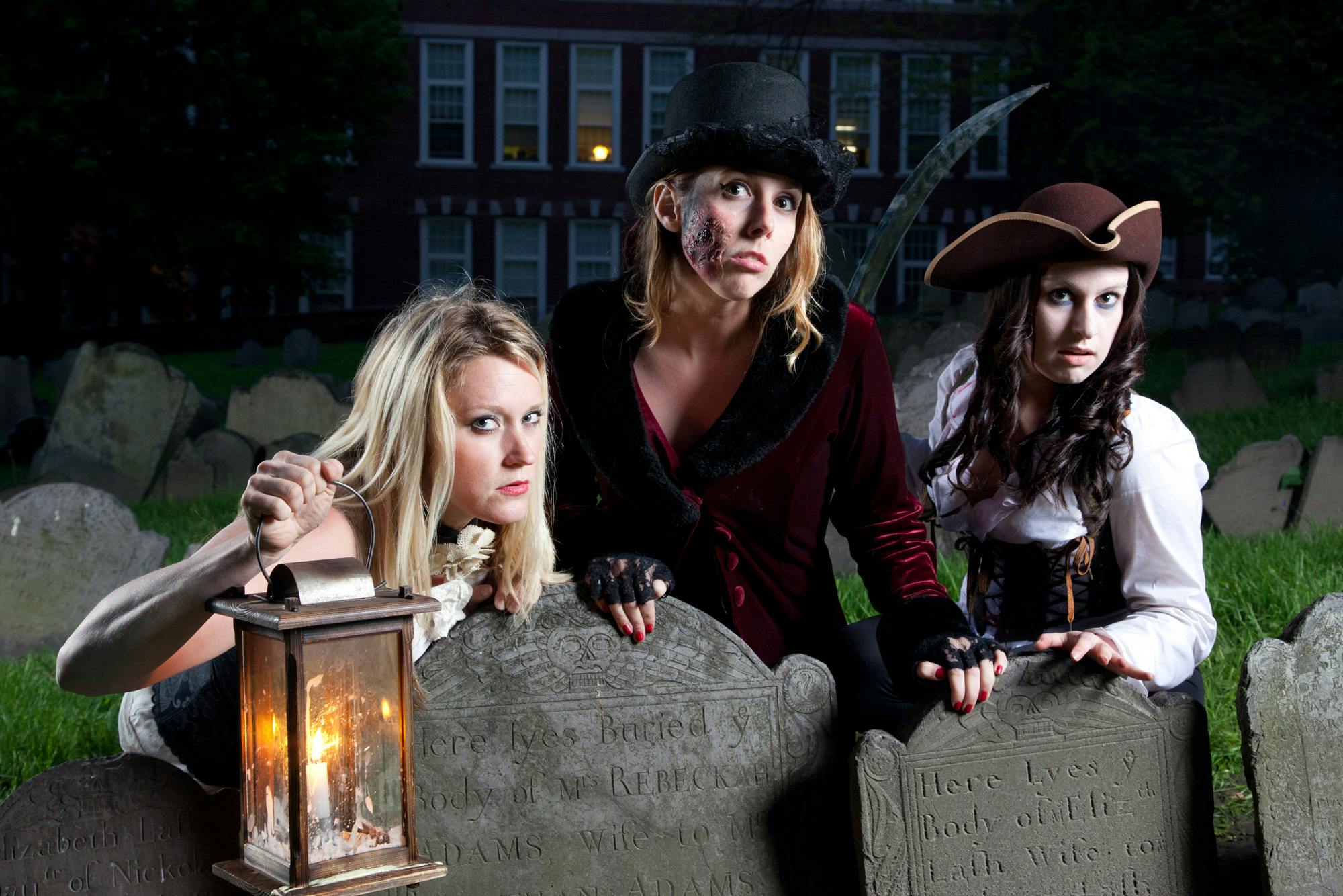 Three women in costume stand in a graveyard at night, positioned behind old tombstones, with a lantern visible on the left.