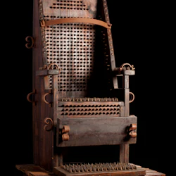 En tortyrstol i trä med många metallspikar på sätet, ryggstödet och armstöden mot en svart bakgrund.