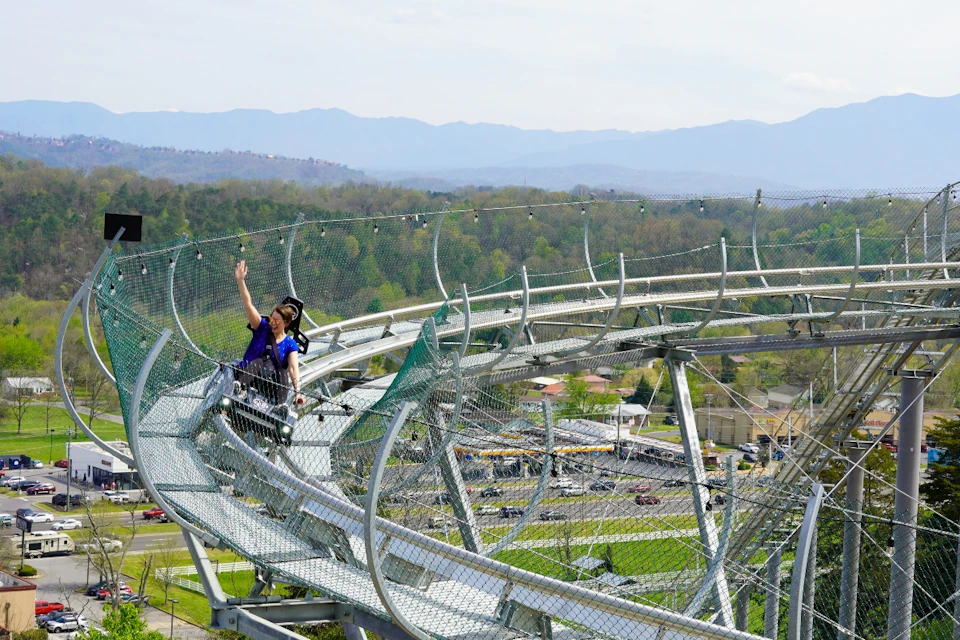 Person waving while riding a scenic mountain roller coaster with a view of distant hills and a village below.