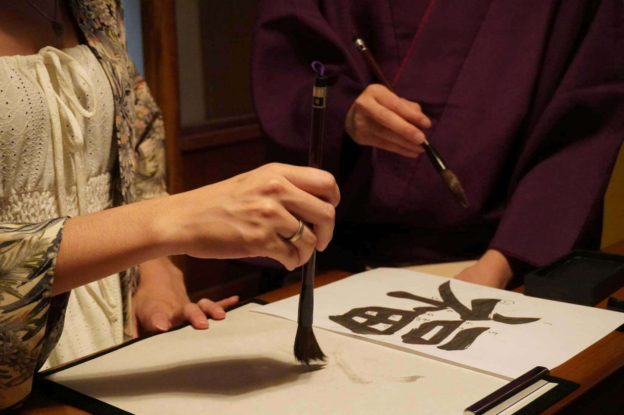 Two people using brushes to create calligraphy on a sheet of paper with visible kanji characters.