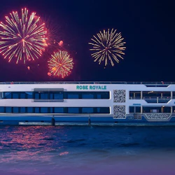 A cruise ship named 'Rose Royale' on water at night with colorful fireworks in the sky above.