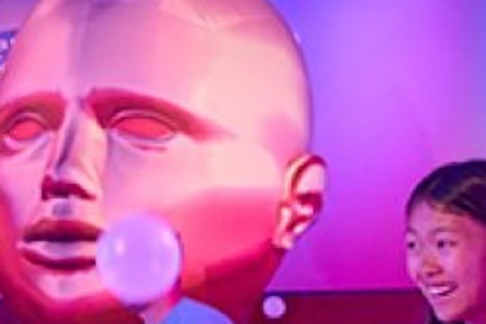 Three people standing in a large ball pit with illuminated white balls, colorful walls, and large head sculpture in the background.