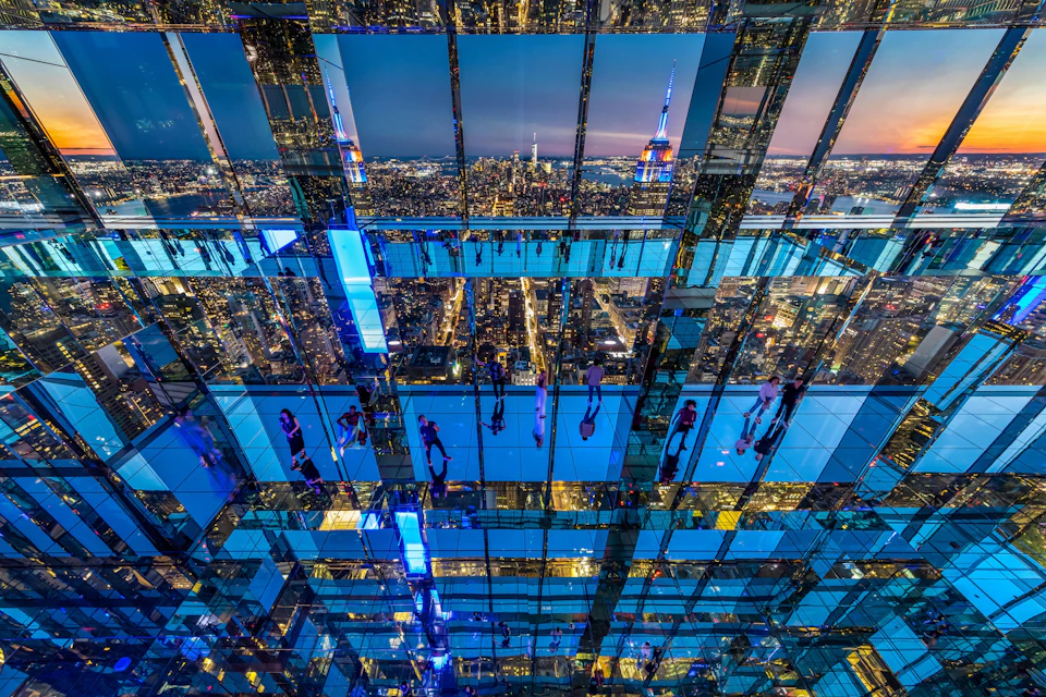 A mirrored interior reflecting people, city lights, and the skyline, including the Empire State Building at dusk.