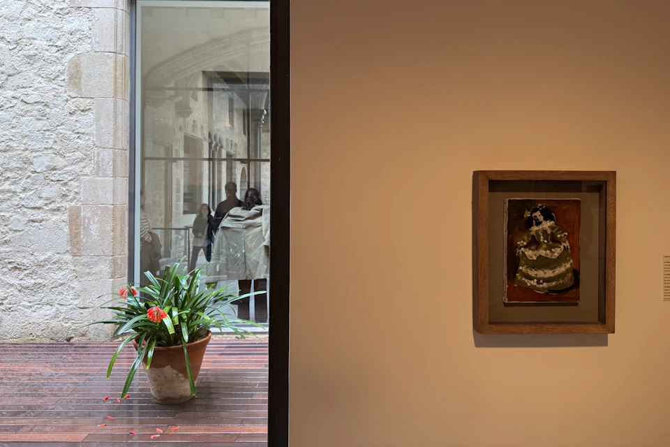 A framed painting of a menina is displayed on a wall, while a large window reflects the museum’s historic courtyard.