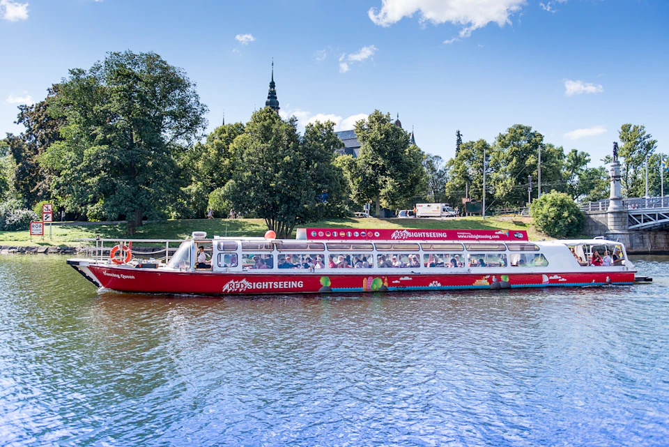 A red sightseeing boat filled with passengers on a river with lush green trees and historic buildings in the background.