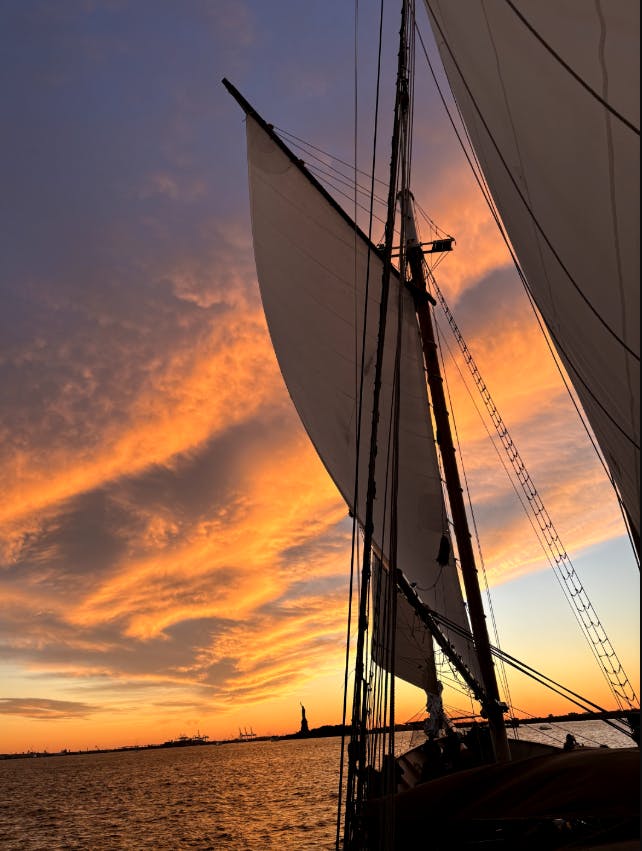 Ένα ιστιοφόρο με μεγάλα πανιά στο ηλιοβασίλεμα, με το Άγαλμα της Ελευθερίας ορατό στο βάθος κάτω από έναν ζωντανό πορτοκαλί ουρανό.