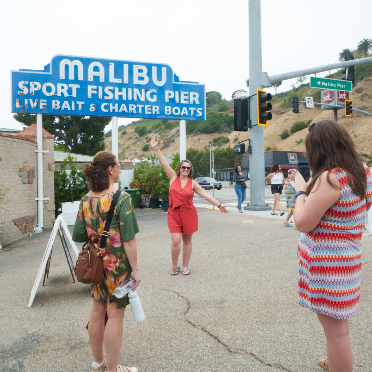 Three women near the Malibu Sport Fishing Pier sign. One poses for a photo while another takes the picture.