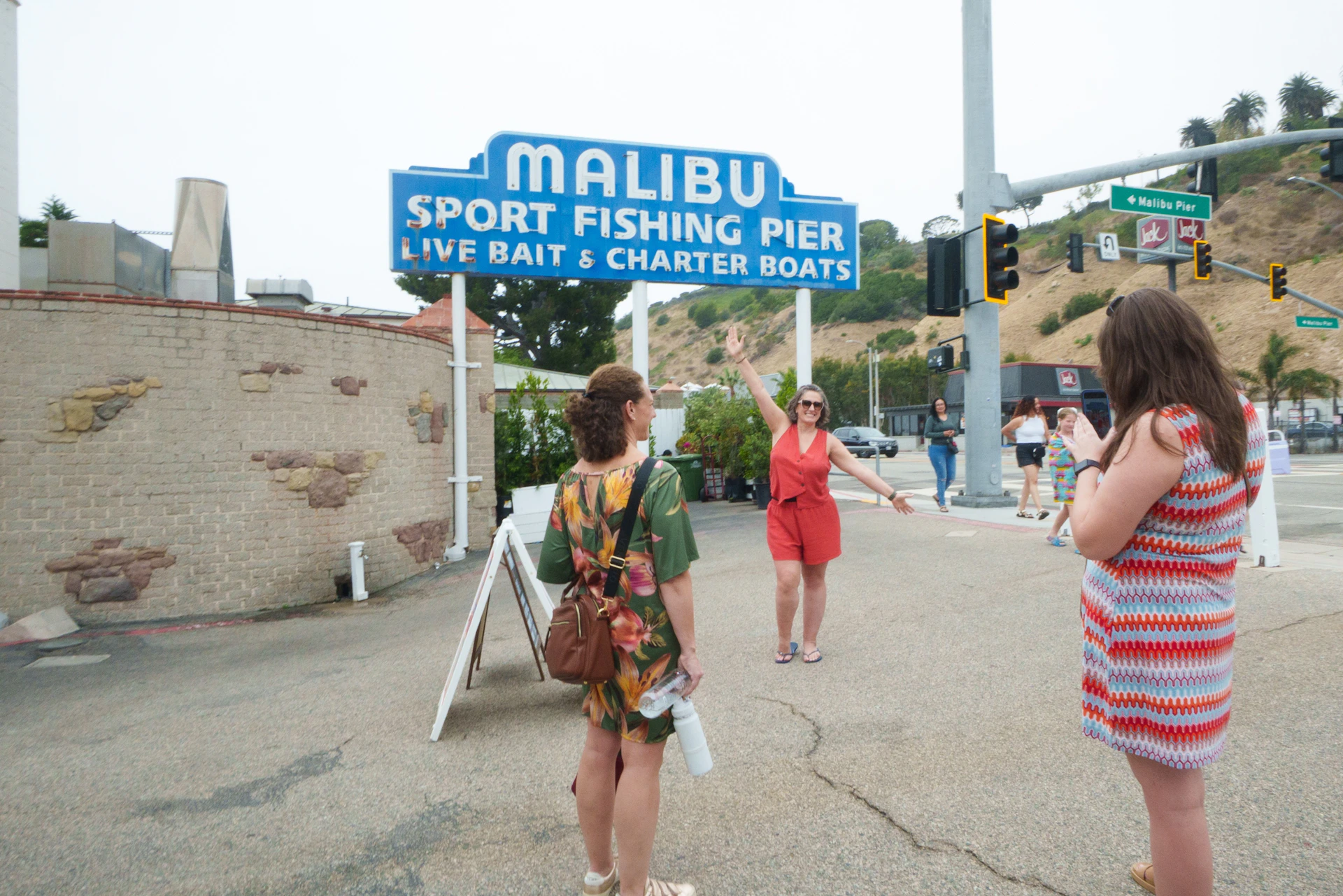 Three women near the Malibu Sport Fishing Pier sign. One poses for a photo while another takes the picture.