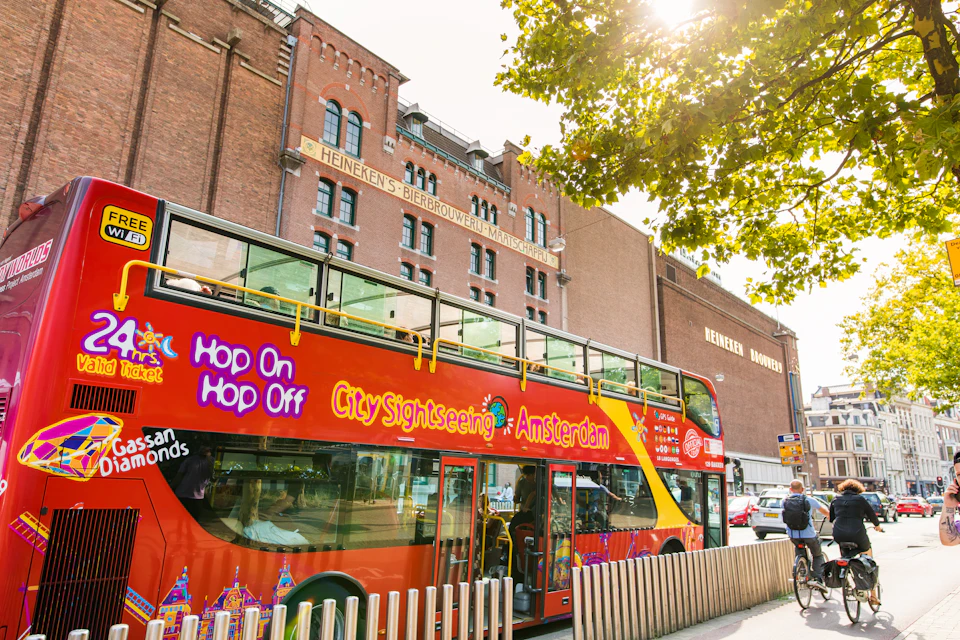 A bright red, double-decker City Sightseeing bus in front of the Heineken brewery building in Amsterdam under sunny skies.