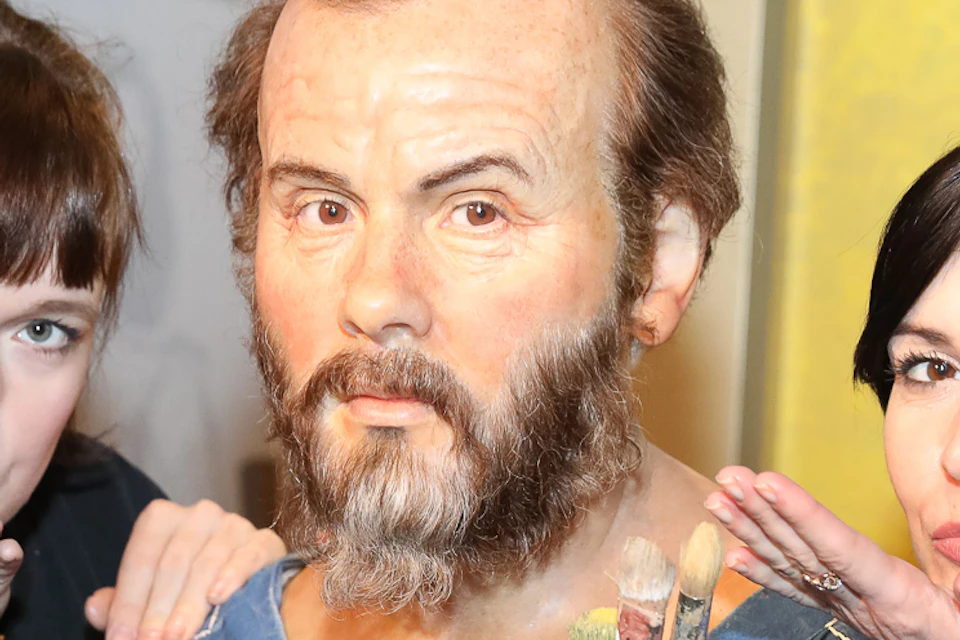 Two women pose next to a bearded wax figure holding paintbrushes, each mimicking a kiss towards the figure's face.
