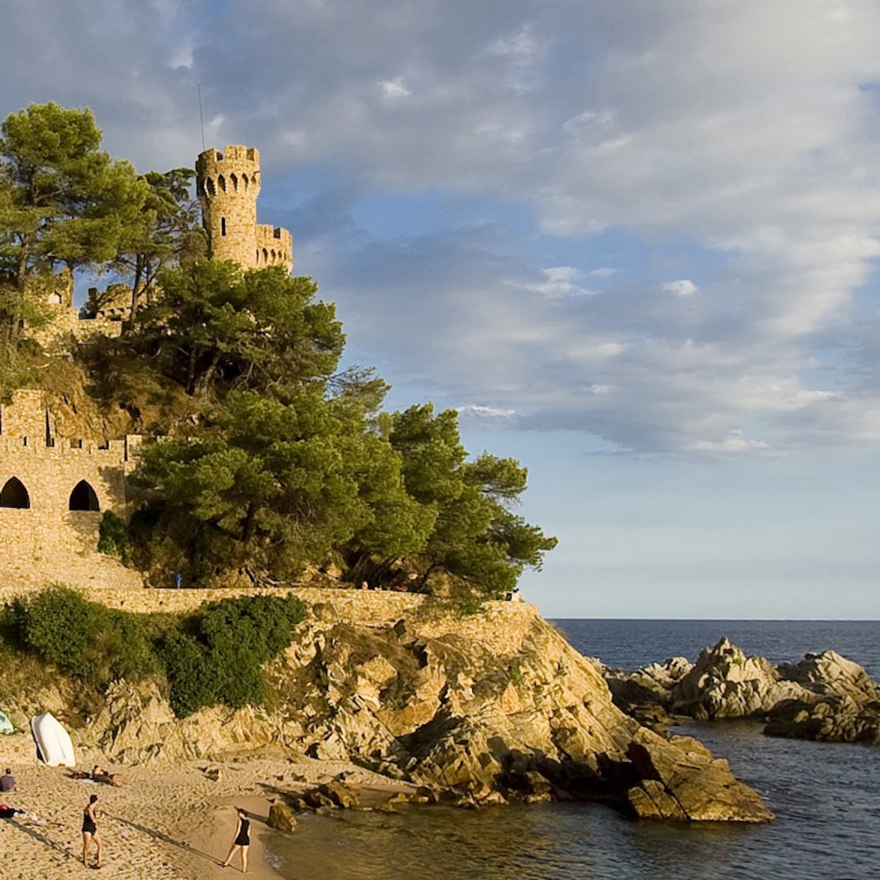Stone castle with tower on a rocky hill beside a beach and ocean. Trees surround the castle. People walk on the sandy shore.