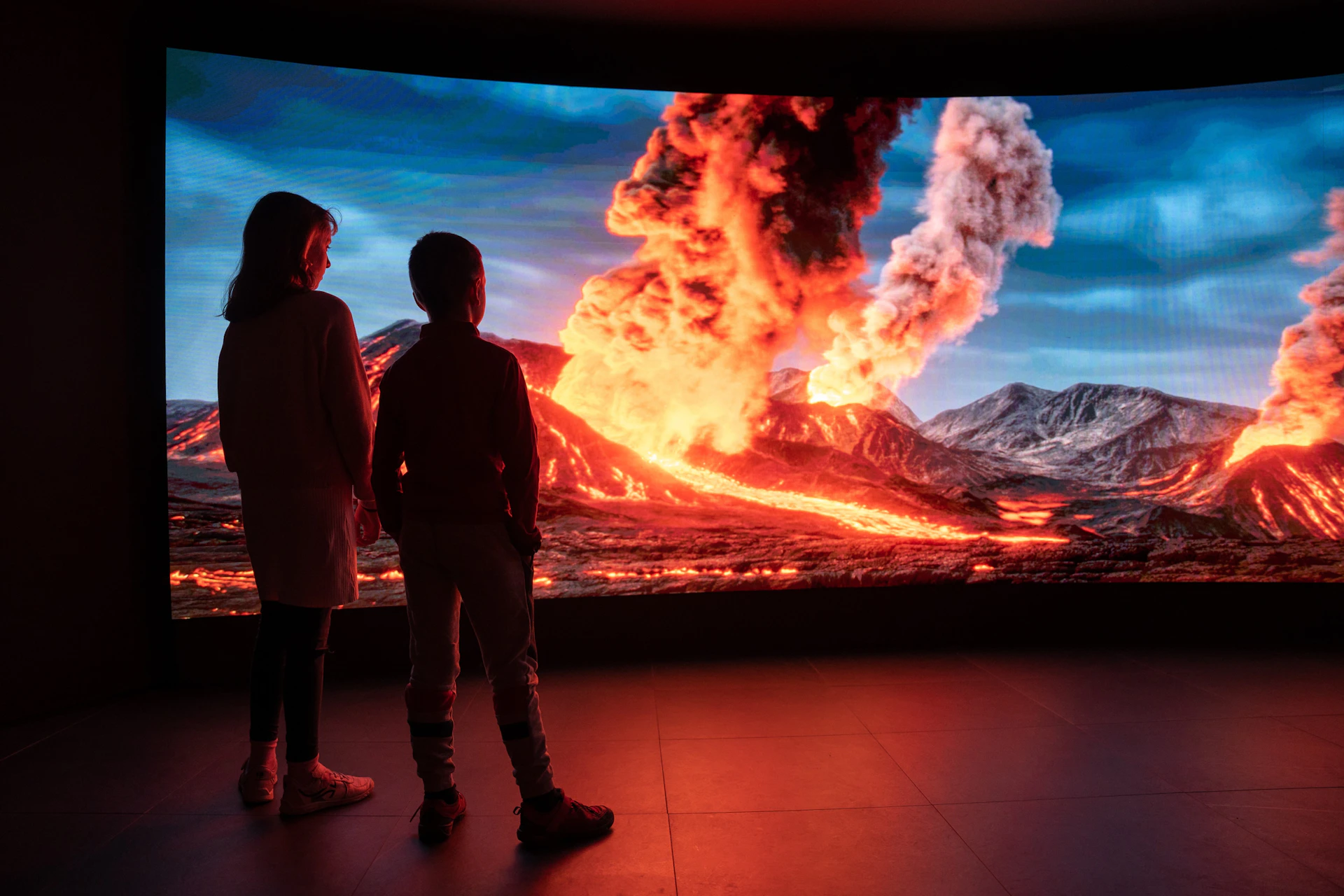 Two children stand in front of a large screen showing a dramatic volcanic eruption with flowing lava and smoke under a blue sky.