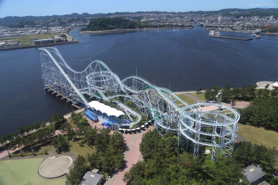 Aerial view of a rollercoaster by a body of water with surrounding trees and a cityscape in the background.