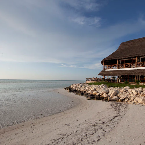 Ein strohgedecktes Gebäude mit Holzbalkonen bietet einen Blick auf den Sandstrand und das ruhige Meer bei klarem Himmel.