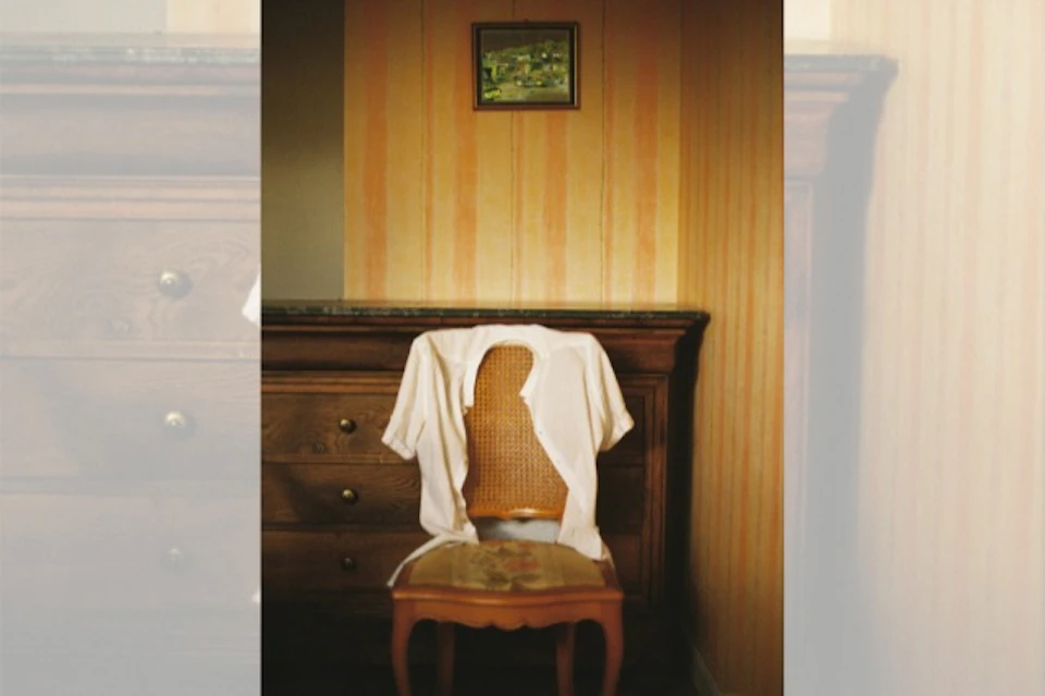 A wooden chair with a white shirt draped over it in front of a wooden chest of drawers, with a framed painting on the wall.