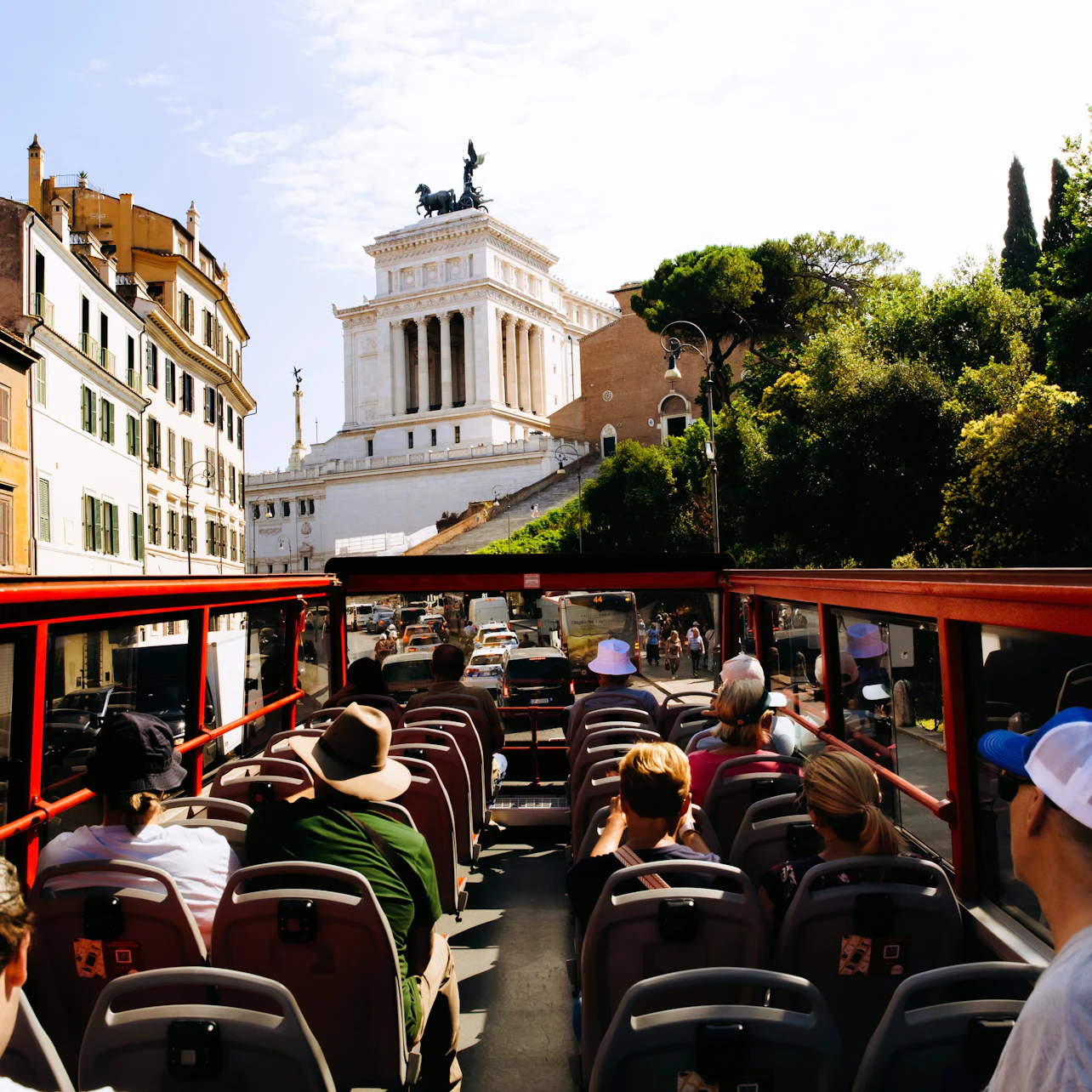 Tourists sitting on the upper deck of a red double-decker bus, viewing historical buildings and monuments in a city.