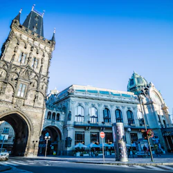 Ett historiskt torn och utsmyckad byggnad med en välvd ingång under en klarblå himmel i en urban miljö.