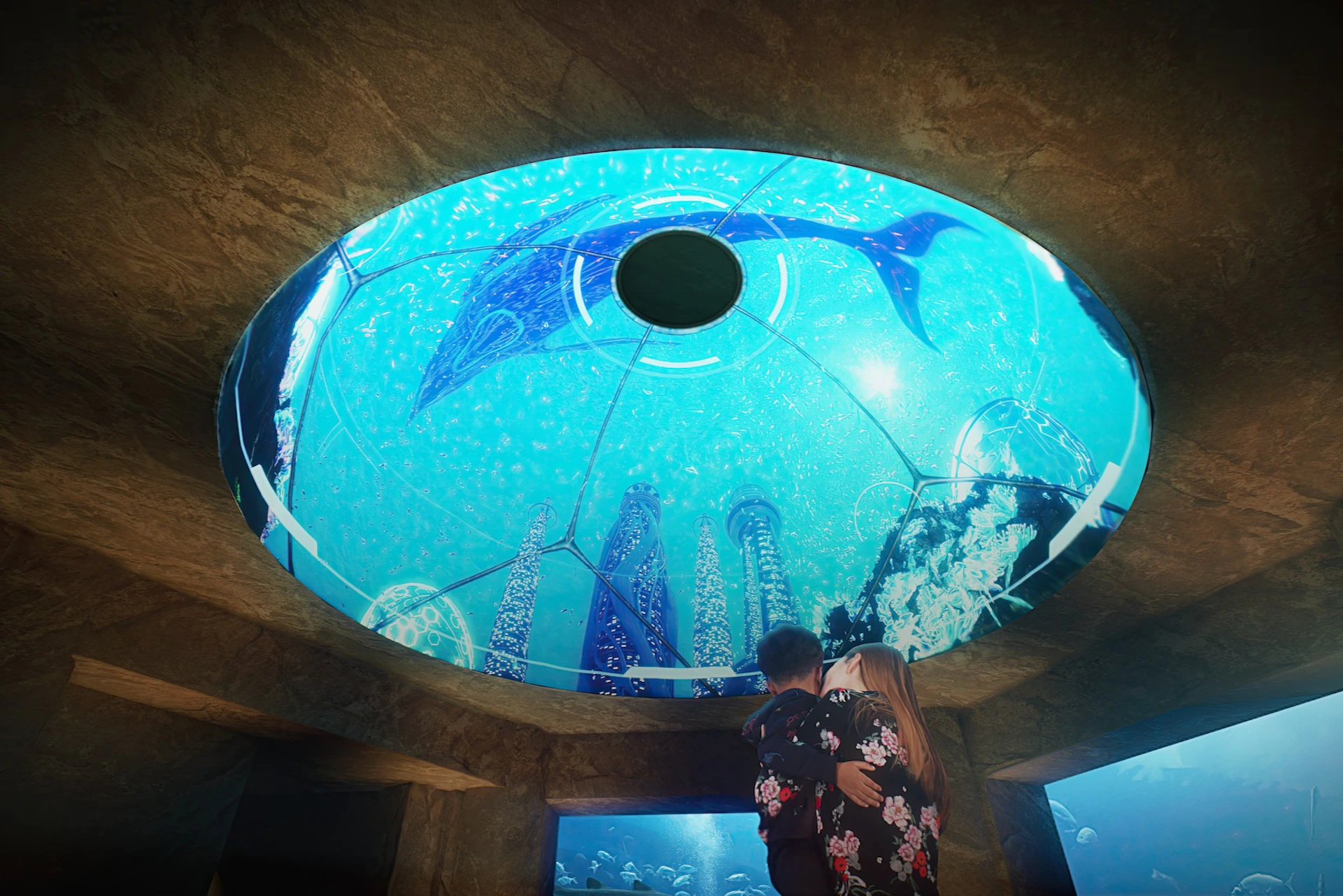 A couple embraces while looking at a large circular aquarium ceiling with underwater structures and a manta ray swimming above.