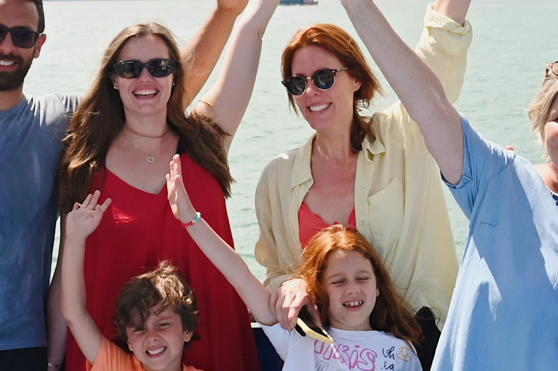 Group of seven people waving and smiling on a boat with a city skyline in the background.