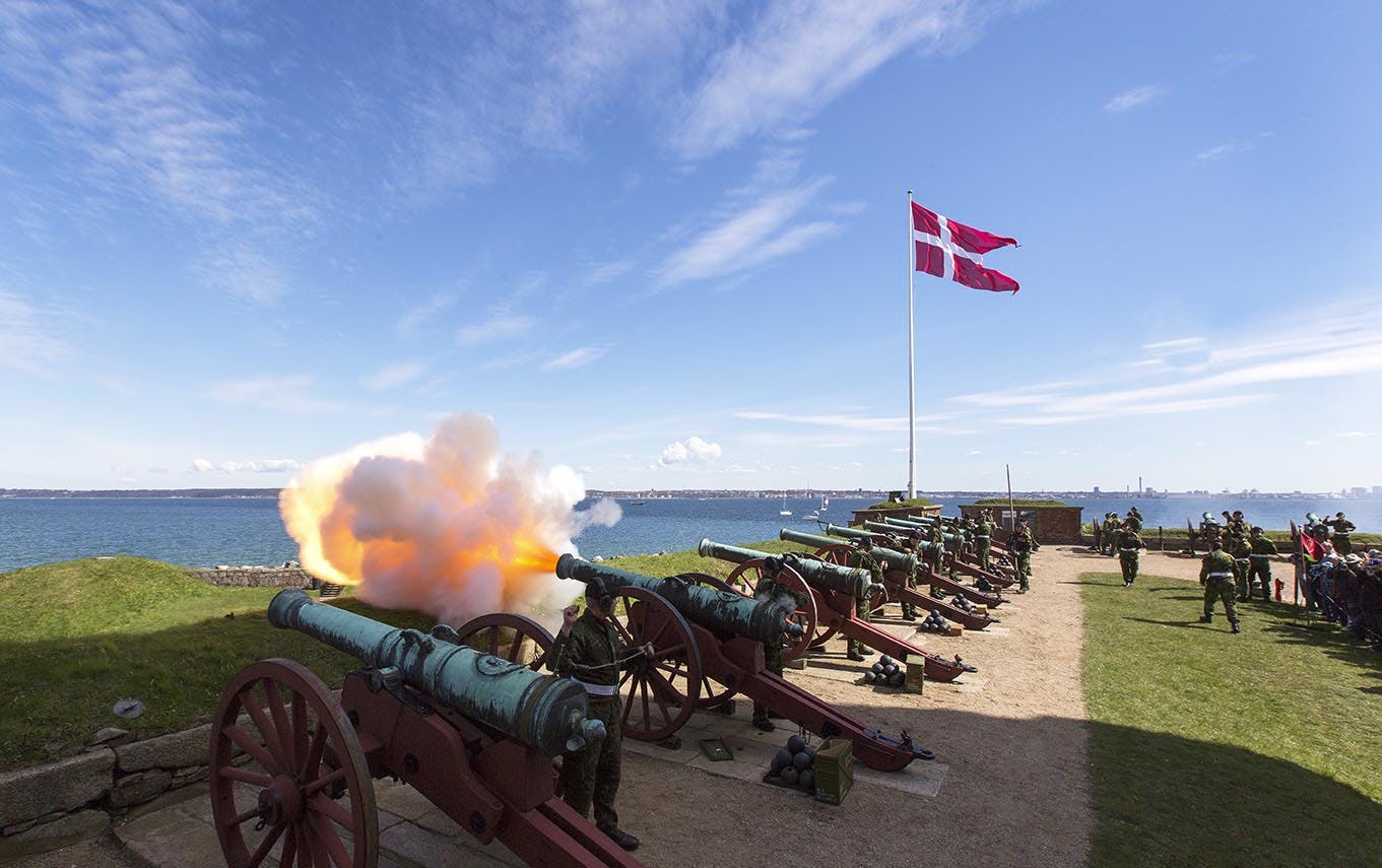 Cannons firing near a coastline with a Danish flag flying on a tall flagpole in the background under a clear sky.