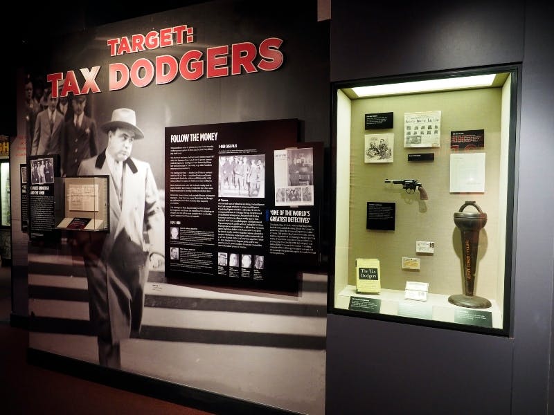 Exhibit display with a black-and-white photo of a man in a suit, text "TARGET: TAX DODGERS," and historical artifacts in a glass case.