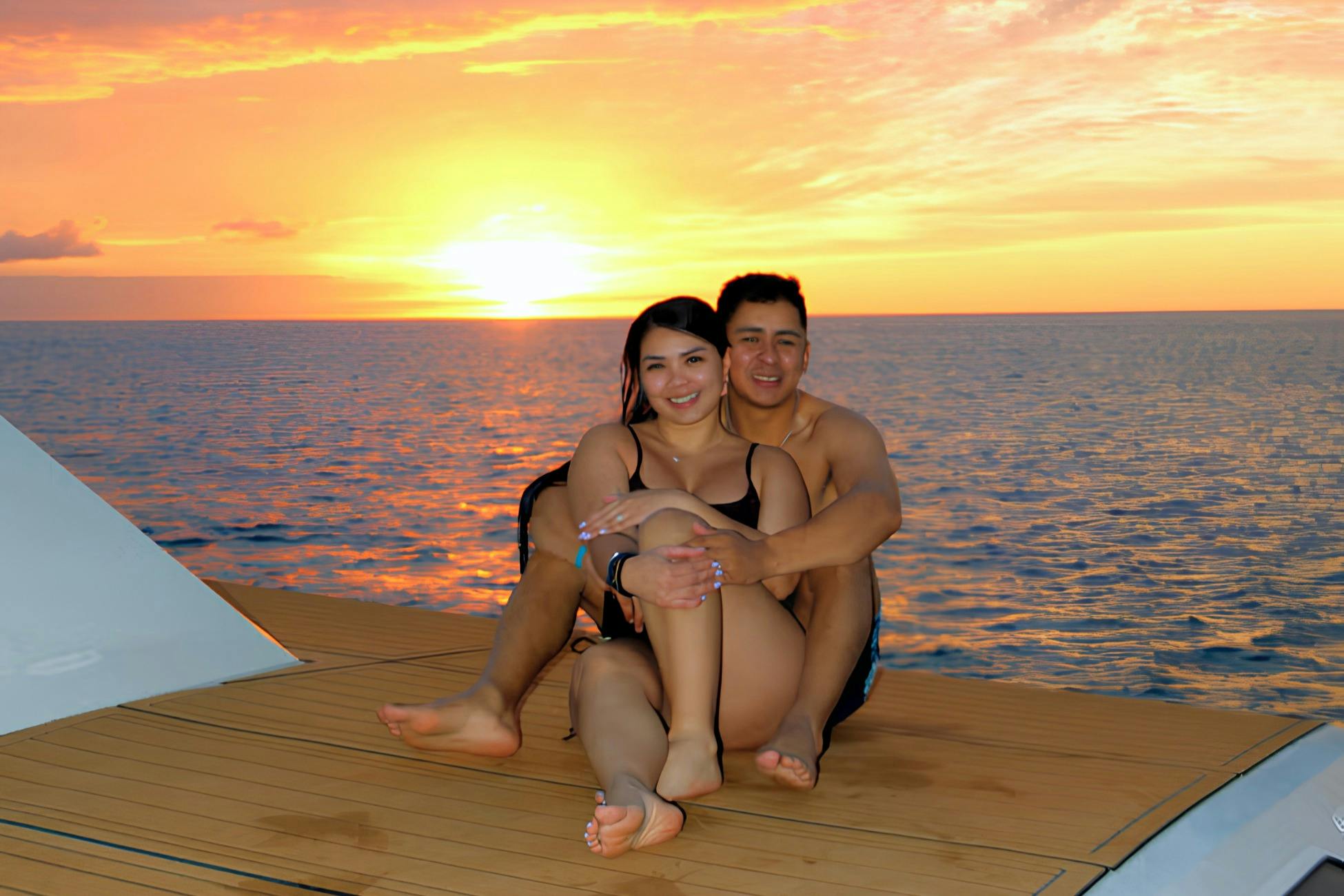 A couple sitting on a wooden deck by the sea at sunset, smiling and embracing each other.
