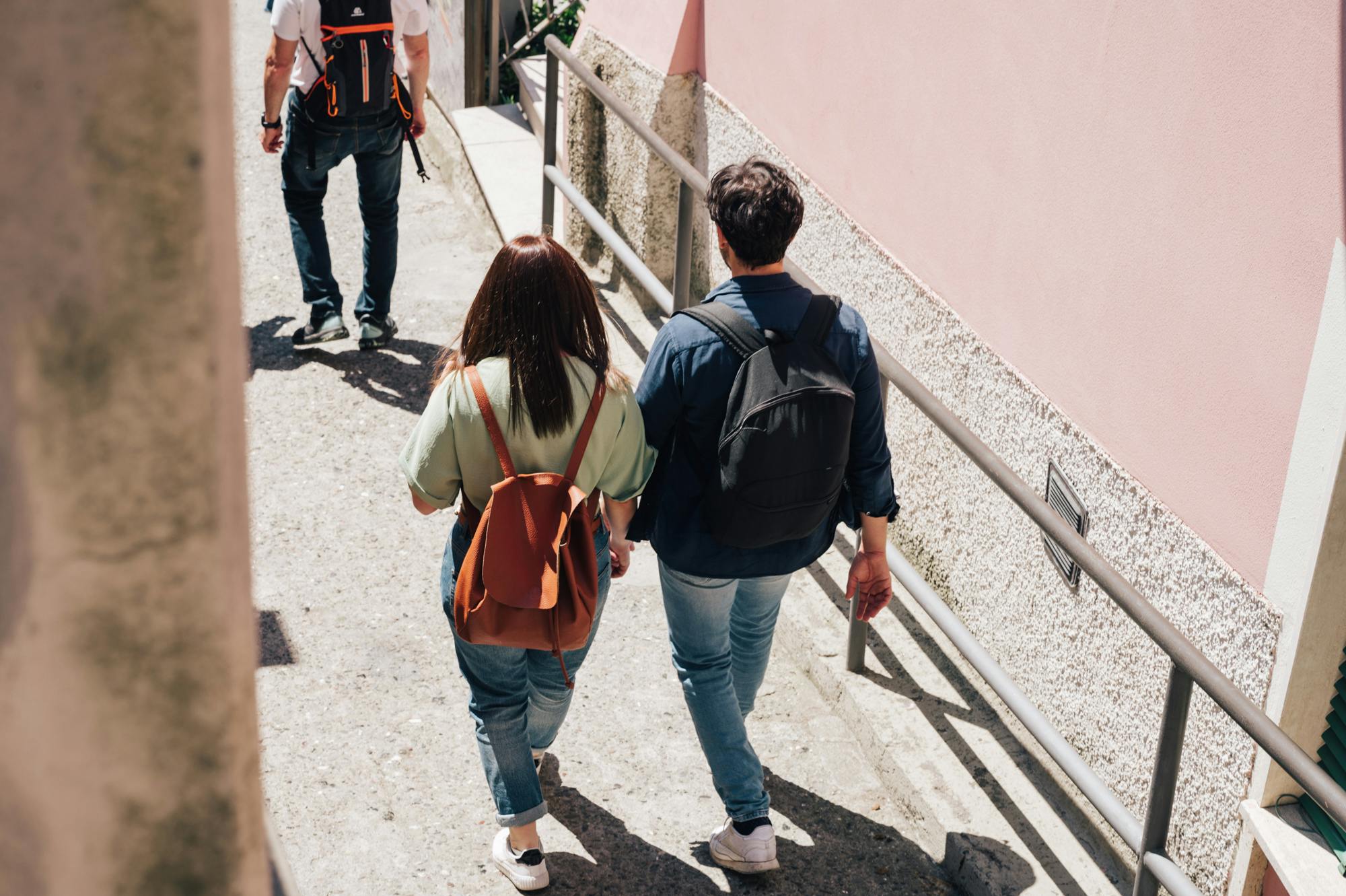 两个背着背包的人走在一条有栏杆的狭窄街道上，后面跟着另一个人。 右侧可见粉红色墙壁。