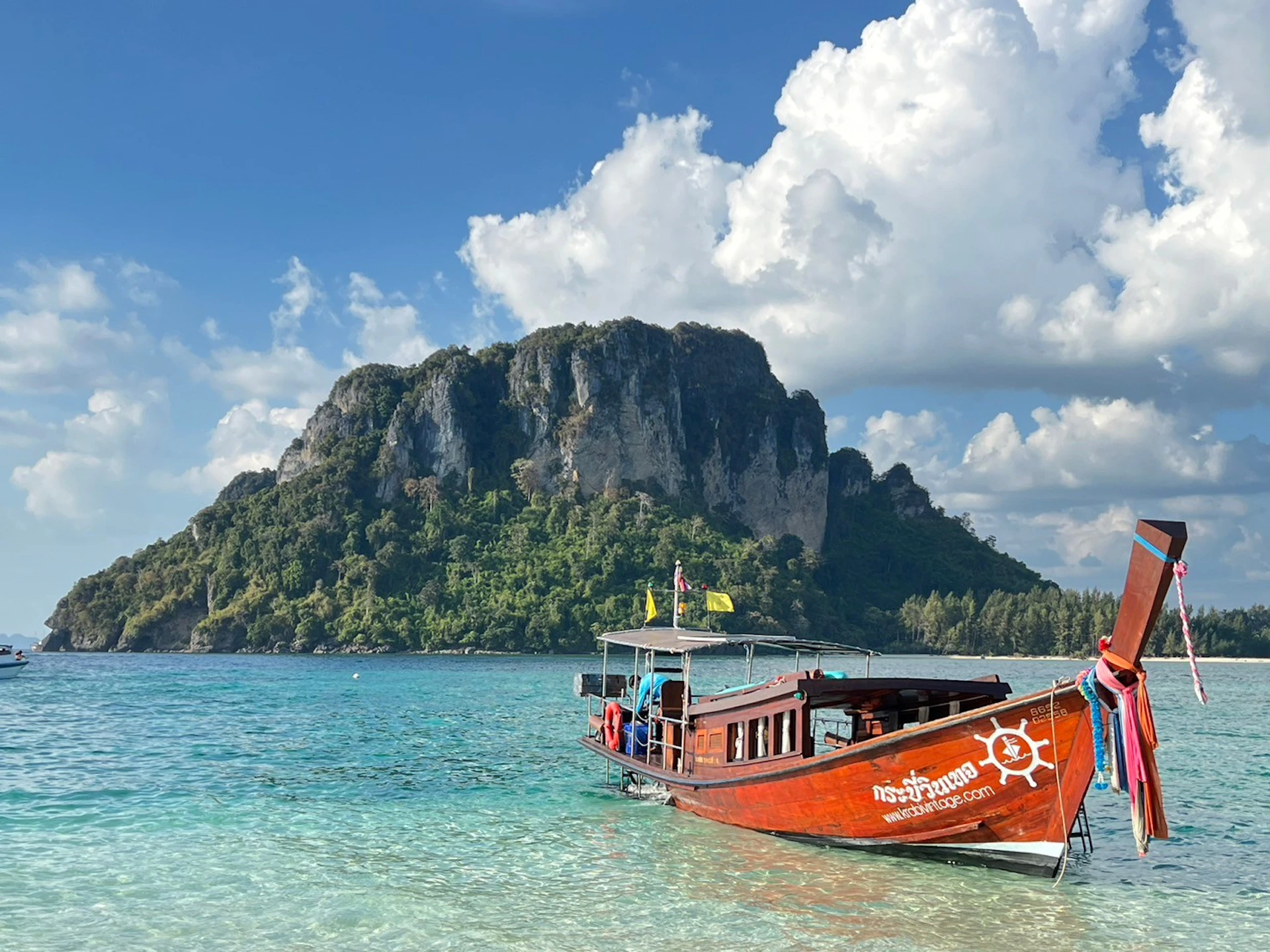 A wooden boat with Thai text is docked in clear turquoise waters, with a large rocky island and cloudy blue sky in the background.