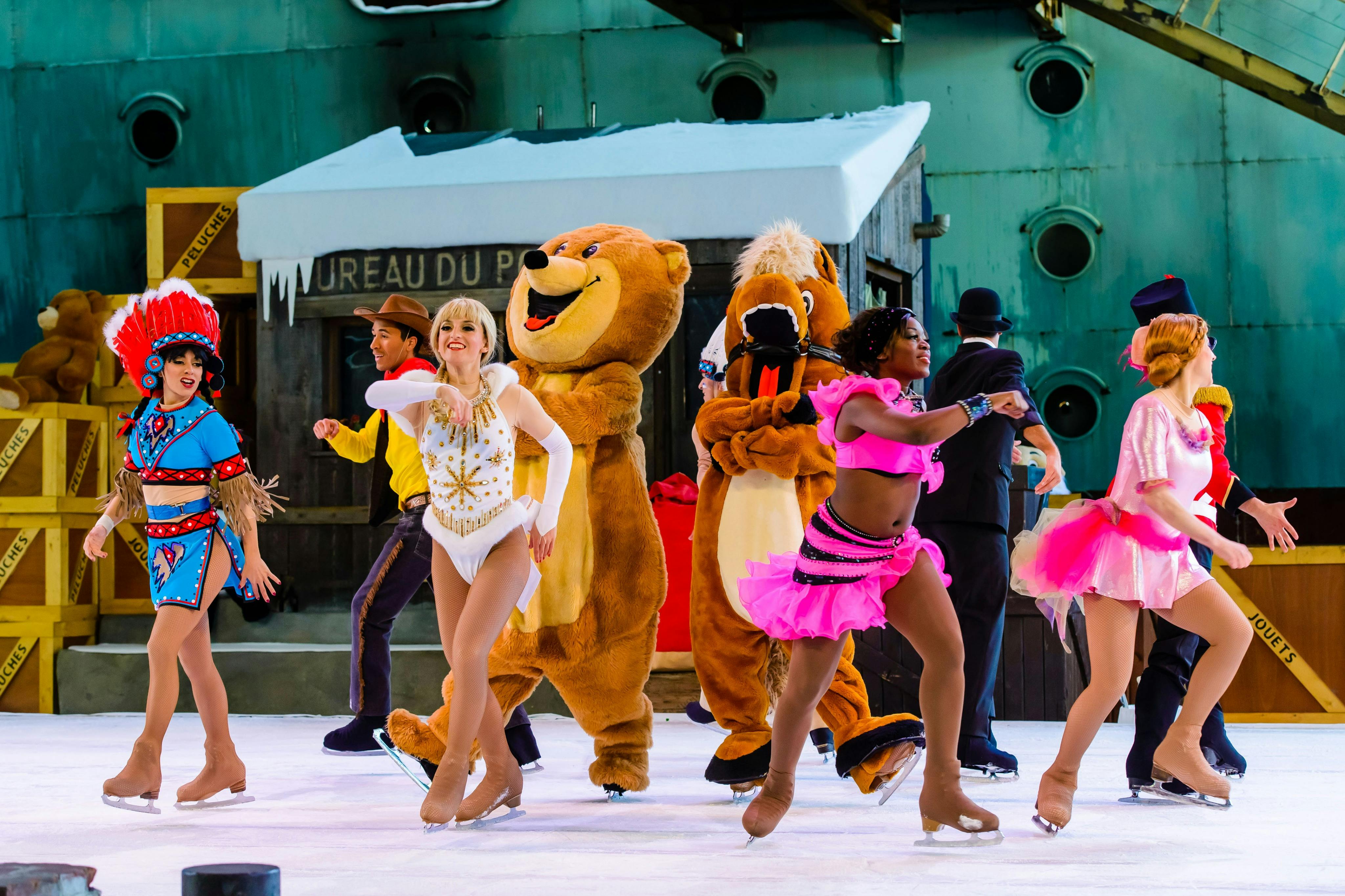 Performers ice skating in colorful costumes, including animal mascots and dancers, on a themed stage with props.
