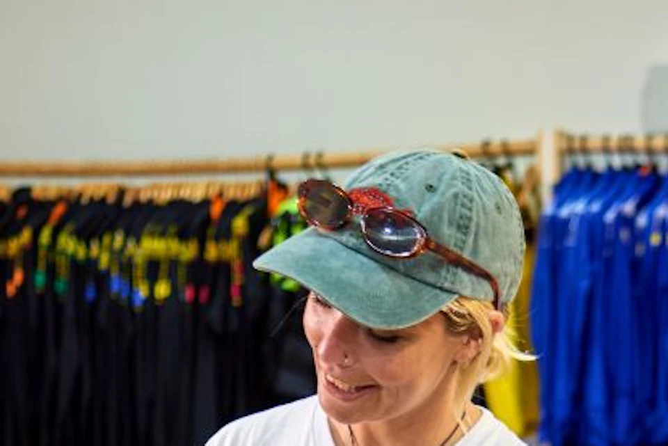 A person in a white shirt and cap showing an electronic tablet to two women in a store with racks of wetsuits in the background.