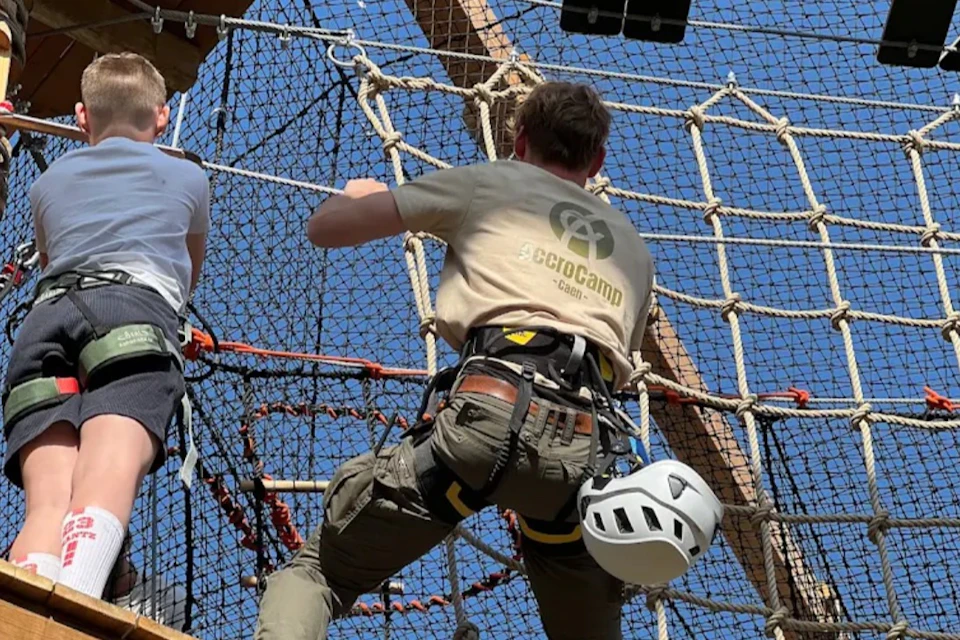Person in a harness climbing a rope net at an outdoor adventure park, with a helmet attached to their belt.