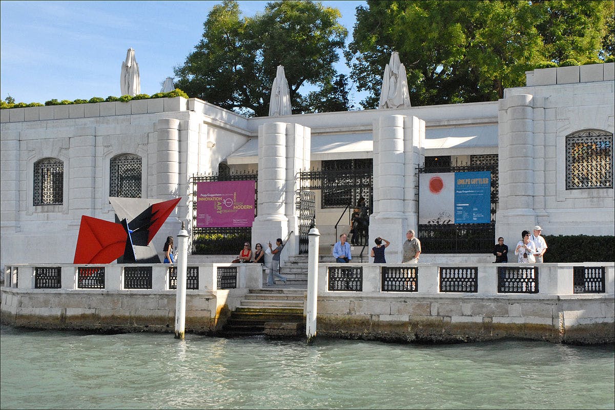 A waterfront museum entrance with people standing outside, two large statues, a colorful abstract sculpture, and informational banners.