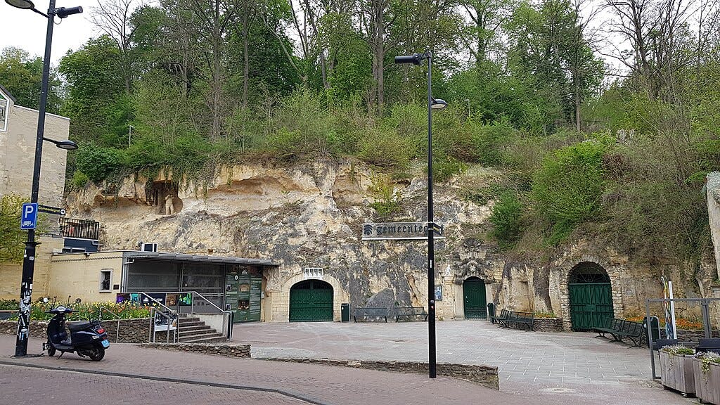 A rocky hillside with green foliage, an entrance labeled "Cementerio," benches, a bike, and a small building in an urban plaza.